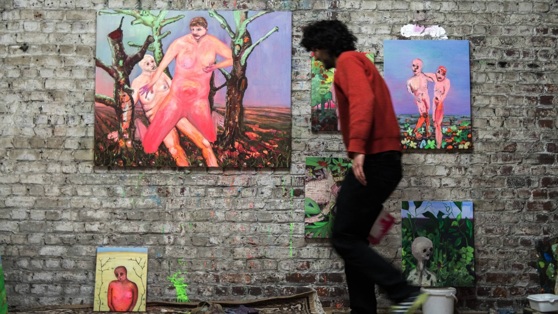 Andriu Deplazes trägt ein Bild zu einer Backsteinwand in seinem Atelier, wo bereits mehrere Bilder aufgehängt sind. Die Bilder hat er mit Ölfarbe aus leuchtenden Farbtönen gemalt.