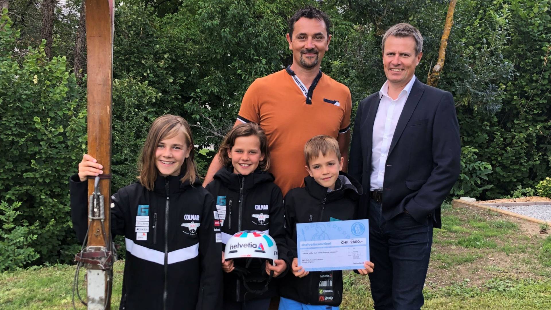  The president of the ski club and his Helvetia customer advisor, plus three young athletes of the ski club, pose with an old wooden ski and the cheque.