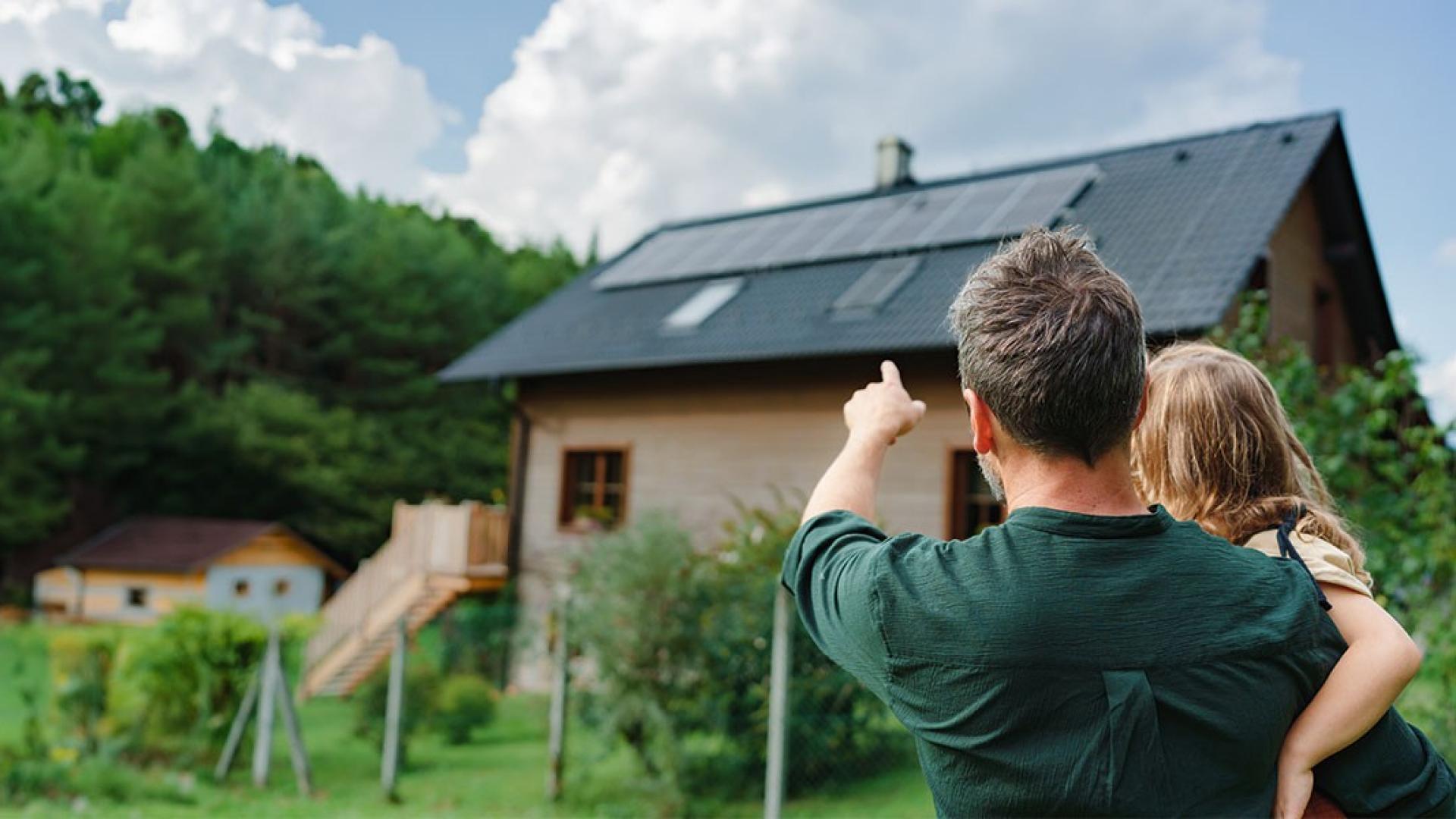 Ein Mann hat ein Kind auf dem Arm und zeigt auf ein Haus, auf dessen Dach eine Photovoltaikanlage montiert ist.