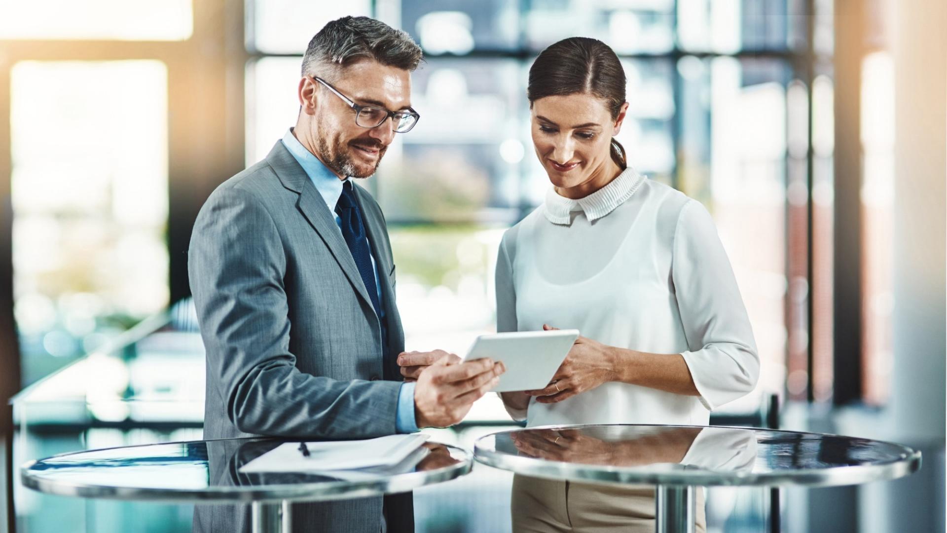 Ein Mann und eine Frau in Business-Kleidung stehen gemeinsam an einem Stehtisch und sehen auf ein Tablet.