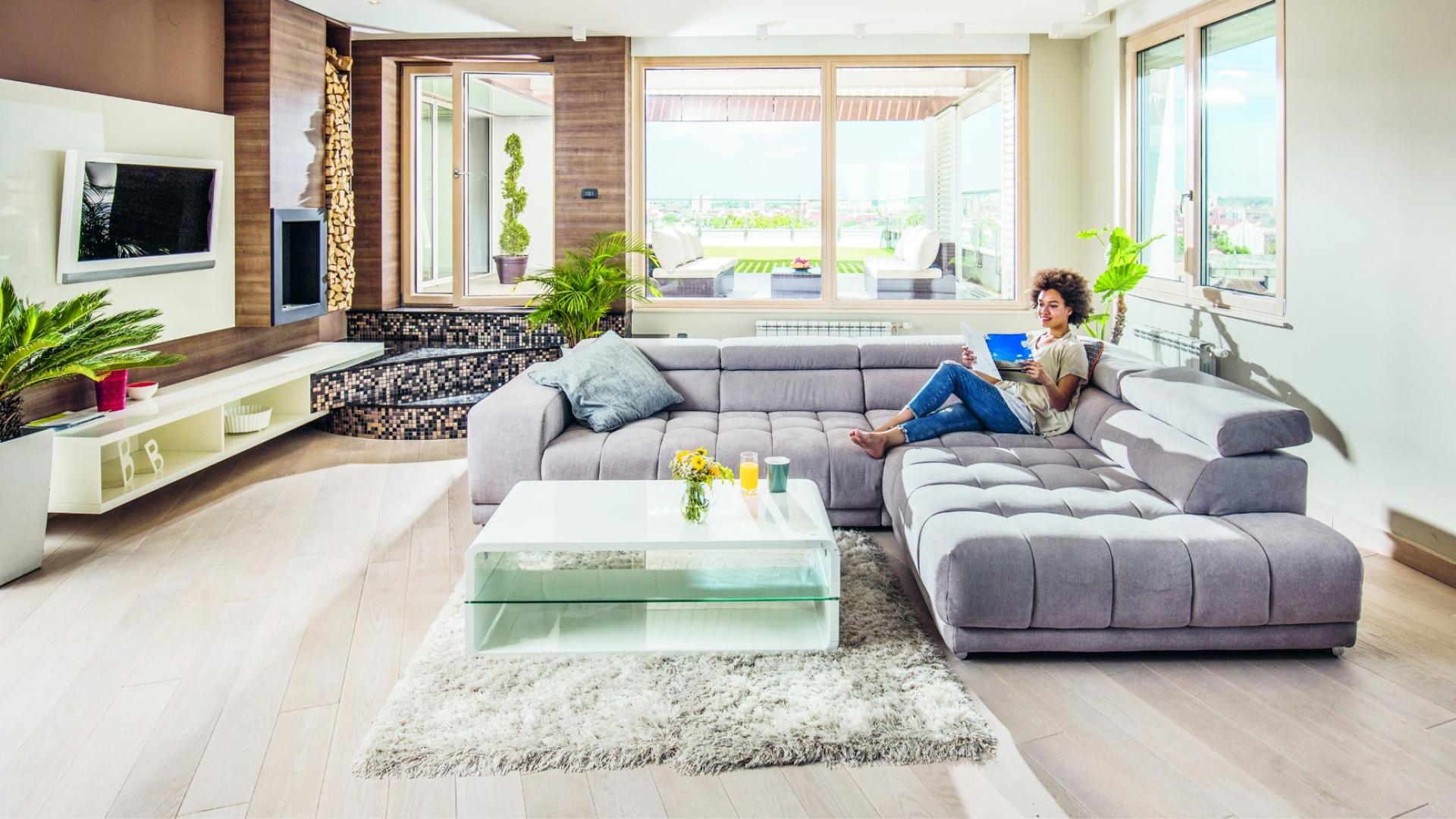 Auf dem Bild ist eine Frau zu sehen, die in ihrem schönen Wohnzimmer auf einer großen, grauen Couch sitzt.