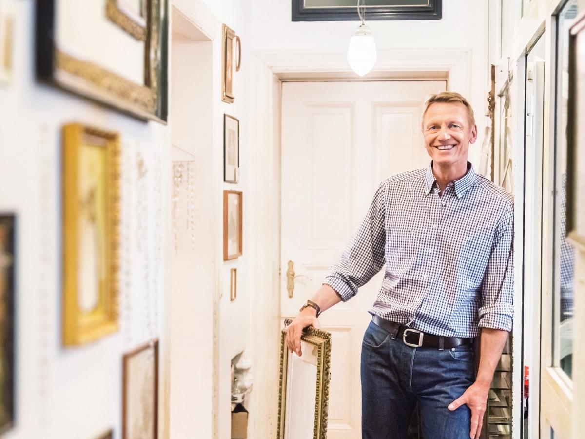Ein erwachsener Mann steht lächelnd in einem Flur, in dem viele Bilder an der Wand hängen, und hält einen Bilderrahmen in der Hand.
