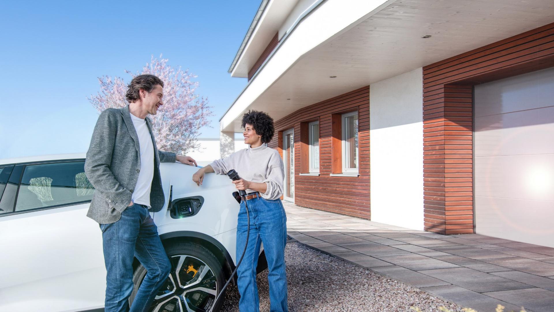 Eine Frau und ein Mann stehen an ein E-Auto angelehnt vor einer Garage.