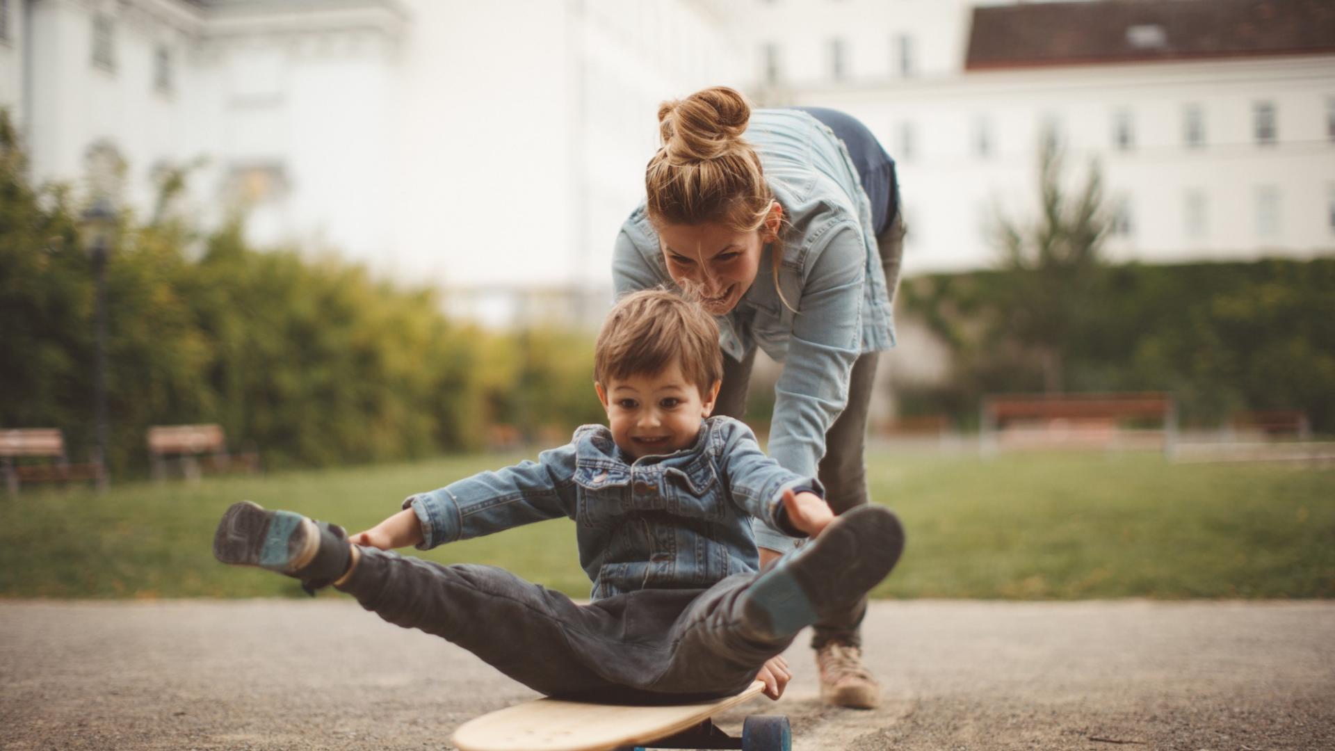 Ein kleiner Junge sitzt auf einem Skateboard und wird von einer Frau angeschubst